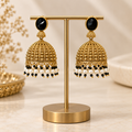 Black and gold brass Jhumka earrings displayed on a metal stand with white and black bead accents