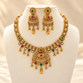 Gold-tone necklace set with red and green accents, ornate central pendant and matching jhumka earrings on a cream bust.