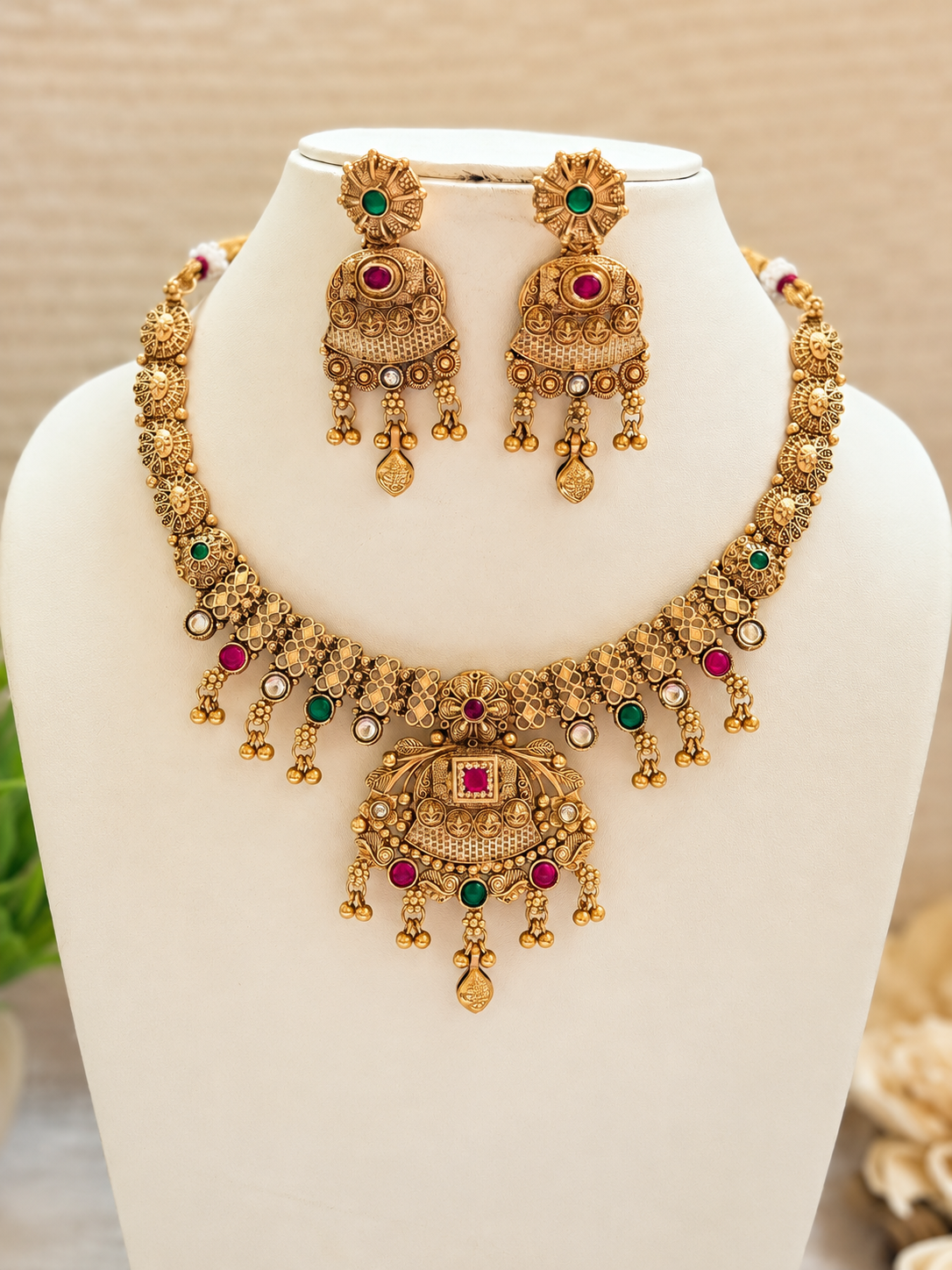 Matching necklace and jhumka earrings displayed on an ivory bust, featuring intricate motifs and red and green accents.